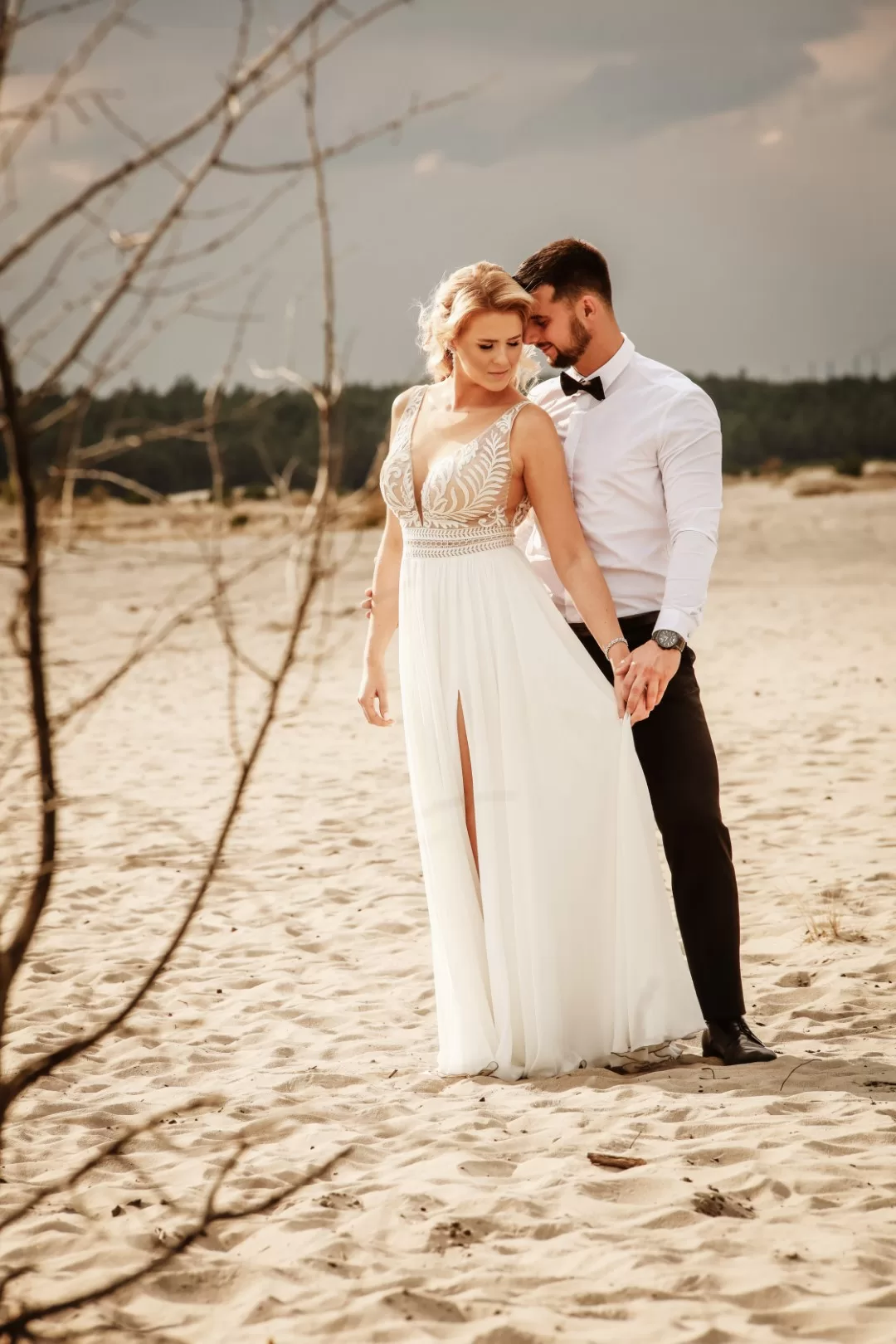 Para młoda na pustyni w surowym klimacie. Delikatnie wtuleni dotykają się głowami. Panna młoda trzyma zwiewną suknię.