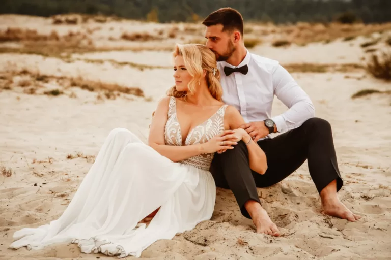 Para młoda siedzi na pustyni, oparci o siebie. Pan młody siedzi boso i całuje w głowę pannę młodą w zwiewnej sukni.