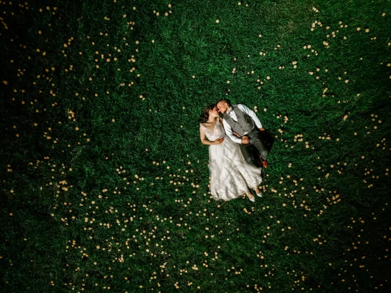 Para młoda podczas pleneru ślubnego, na zdjęciu z drona, leży na łące z kwiatami. Wpatrzeni w siebie trzymają się za ręce.