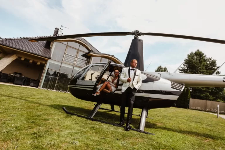 Para młoda pozuje przy helikopterze, przed wylotem na swój ślub. Pan młody w białej marynarce oparty o helikopter, panna młoda siedzi wewnątrz w koronkowej sukni.