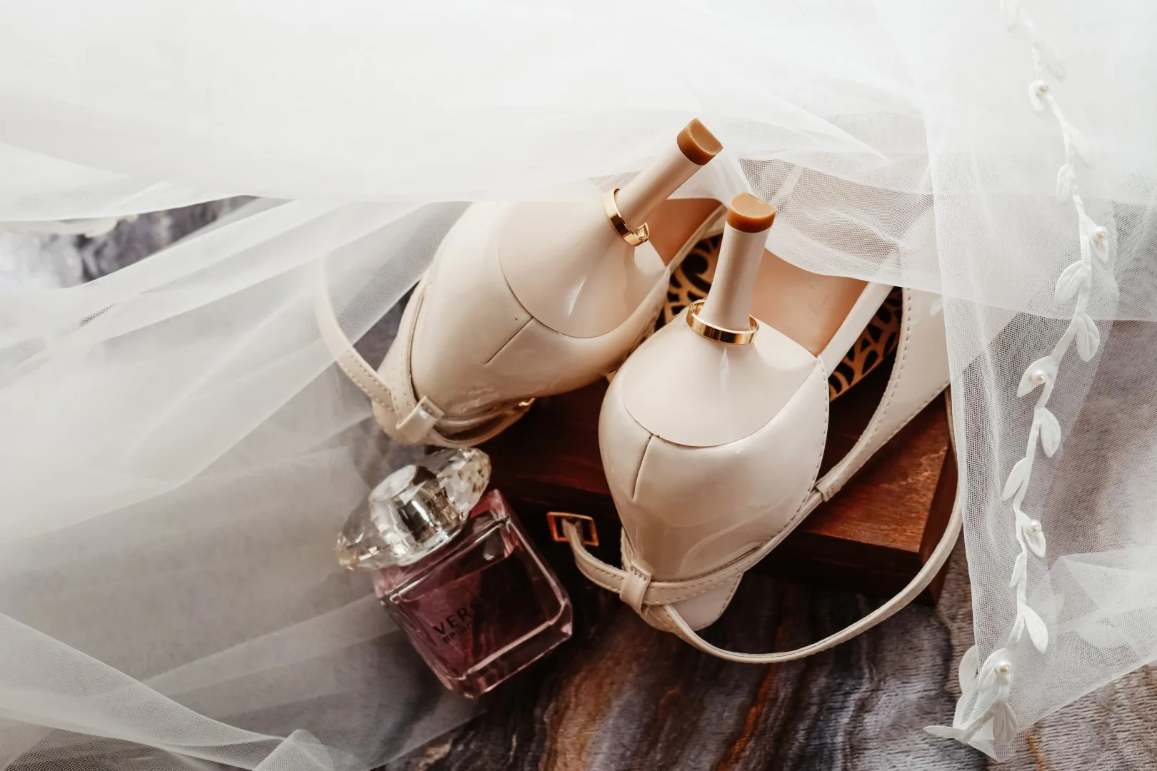 Buty do ślubu w pięknym kremowym kolorze otoczone białym welonem. Złote obrączki zawieszone na obcasach w wymownym zdjęciu przedstawiające detal