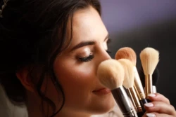 Fotografia śubna Panna młoda z naturalnym makijażem zamyka oczy przykładając do twarzy pędzle.